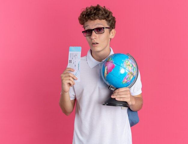 Student guy in casual clothing wearing glasses with backpack holding globe and air ticket looking at camera confused standing over pink background