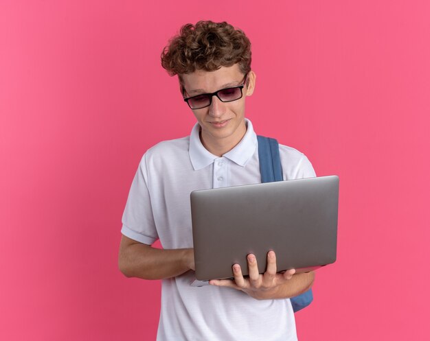 ピンクの背景の上に立っている顔に笑顔でそれを見てラップトップを保持しているバックパックと眼鏡をかけてカジュアルな服を着た学生の男