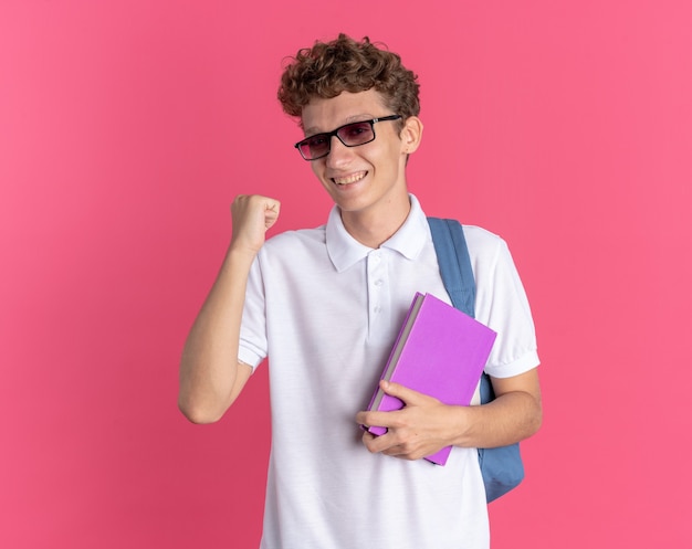 Student guy in casual clothing wearing glasses with backpack holding notebook clenching fist