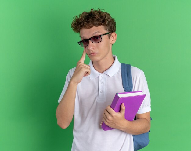 Student guy in casual clothing wearing glasses with backpack holding notebook looking at camera pointing at his eye with index finger standing over green background