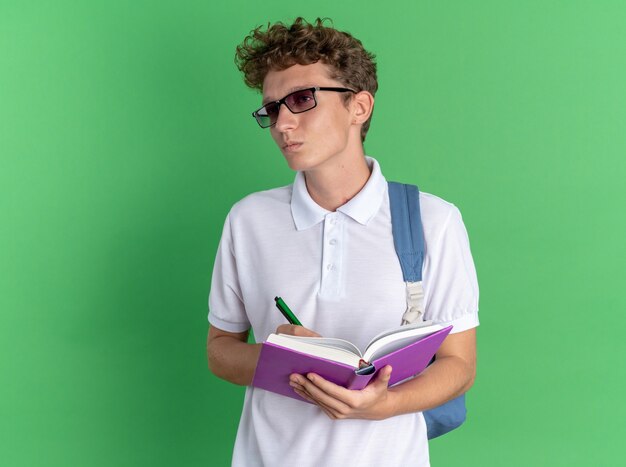 Student guy in casual clothing wearing glasses with backpack holding notebook and pen writing something with serious face