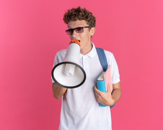 Student guy in casual clothing wearing glasses with backpack holding notebooks shouting to megaphone standing over pink background