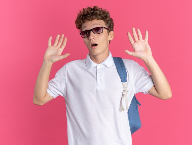 Student guy in casual clothing wearing glasses with backpack raising arms looking confused standing over pink background