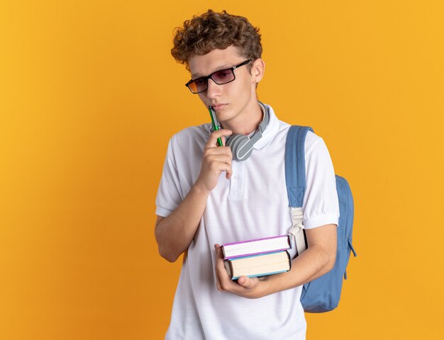 Student guy in casual clothing with headphones wearing glasses with backpack holding books looking puzzled standing over orange background