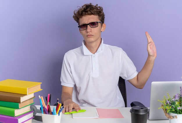 흰색 폴로 셔츠를 입은 학생 남자가 책상에 앉아 책을 들고 진지한 얼굴로 카메라를 쳐다보며 파란색 배경 위에 손을 들고 방어 제스처를 취합니다.