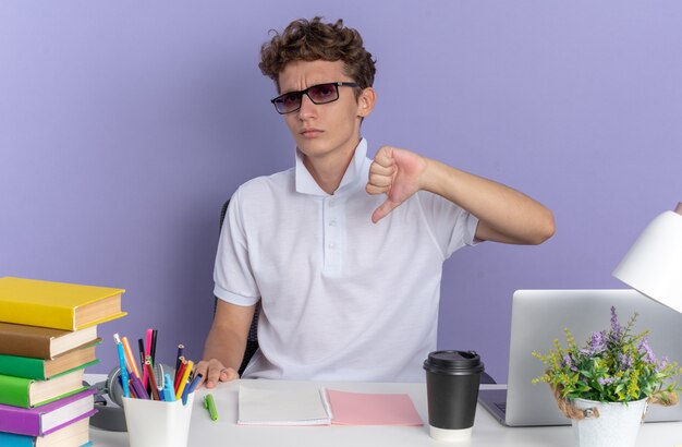 Студент парень в белой рубашке поло в очках сидит за столом с книгами, глядя в камеру с серьезным лицом, указывая на себя на синем фоне