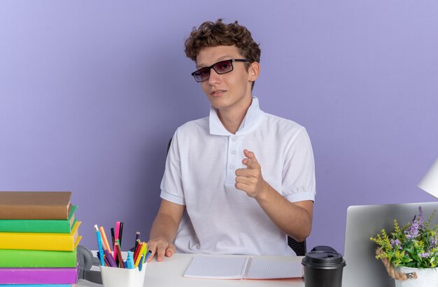 Student guy in white polo shirt wearing glasses sitting at the table with books looking confident pointing with index finger at camera over blue background