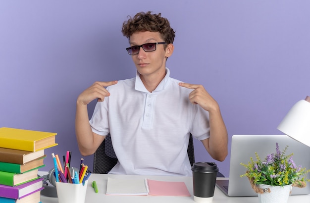 Foto gratuita ragazzo studente in polo bianca con gli occhiali seduto al tavolo con libri dall'aria fiduciosa sorridente che indica se stesso su sfondo blu