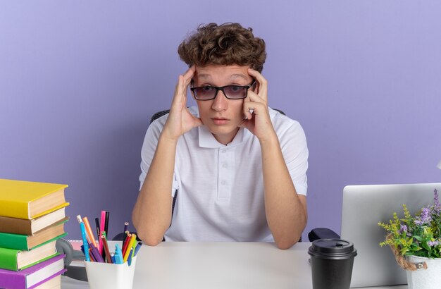 Student guy in white polo shirt wearing glasses sitting at the table with books looking confused over blue background