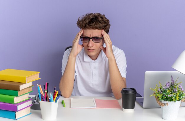 Студент в белой рубашке поло в очках сидит за столом с книгами и выглядит усталым и раздраженным, касаясь своей головы над синей поверхностью