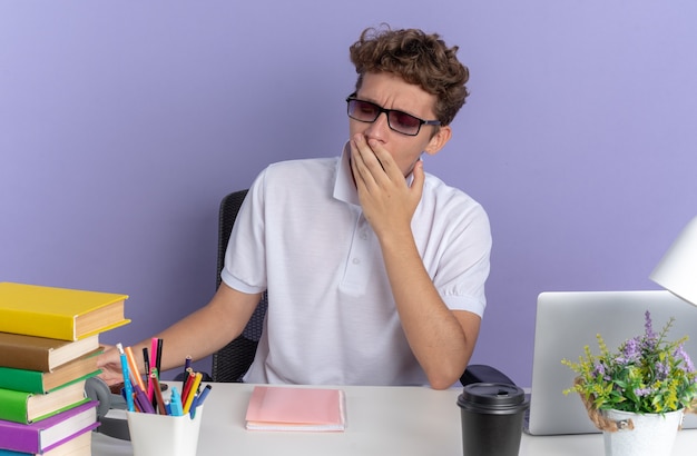 Student guy in white polo shirt wearing glasses sitting at the table with books looking tired and bored yawning over blue surface