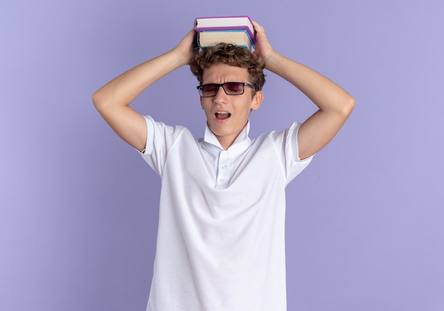 Student guy in white polo shirt and yellow cap wearing glasses holding notebooks over his head looking annoyed and tired standing over blue background