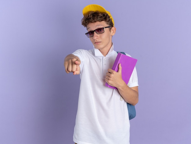 Student guy in white polo shirt and yellow cap wearing glasses with backpack holding notebook pointing with index finger at camera being surprised