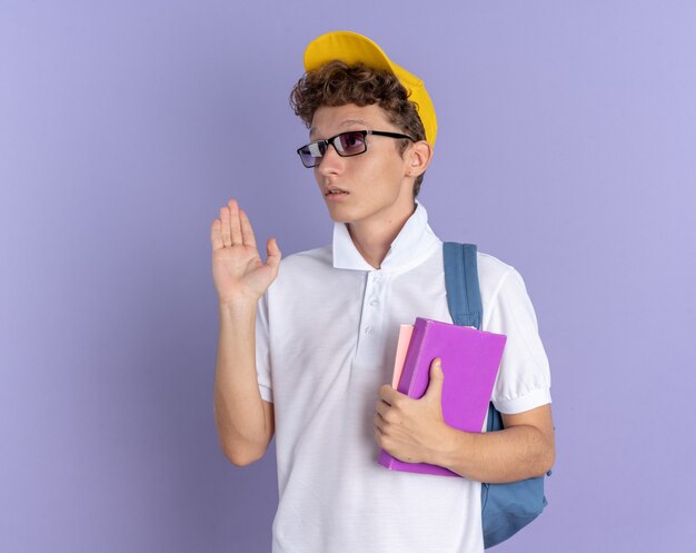 白いポロシャツと黄色の帽子をかぶった学生の男は、青い背景の上に立っている手で手を振って混乱して脇を見てノートを保持しているバックパックと眼鏡をかけています