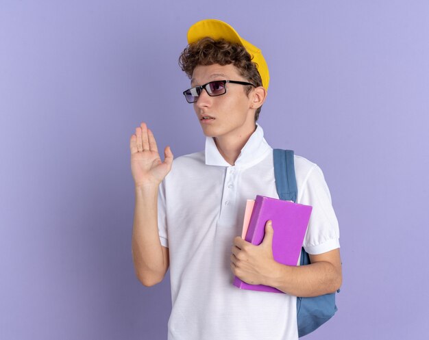 Student guy in white polo shirt and yellow cap wearing glasses with backpack holding notebooks looking aside confused waving with hand standing over blue background