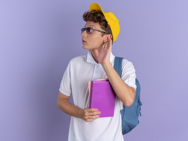 Student guy in white polo shirt and yellow cap wearing glasses with backpack holding notebooks looking aside intrigued with hand over ear trying to listen standing over blue background