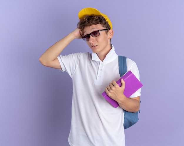 Student guy in white polo shirt and yellow cap wearing glasses with backpack holding notebooks looking at camera confused scratching head standing over blue background