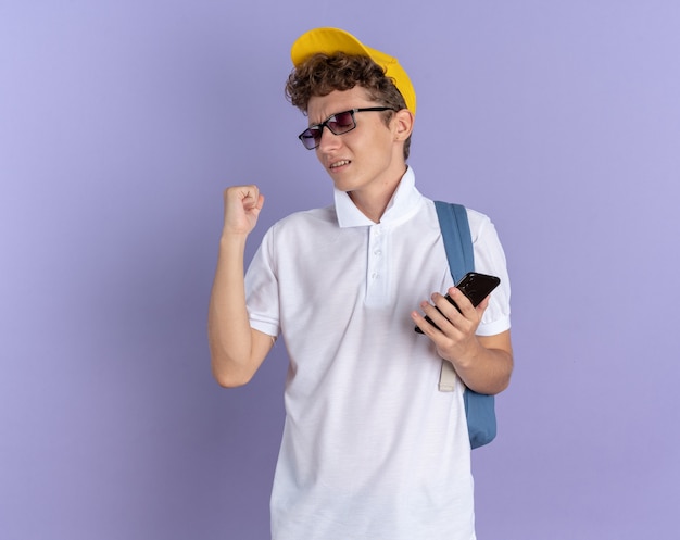 白いポロシャツと黄色い帽子をかぶった学生の男は、青い背景の上に立って彼の成功を喜んで喜んで興奮しているスマートフォンを握りこぶしを握っているバックパックと眼鏡をかけています