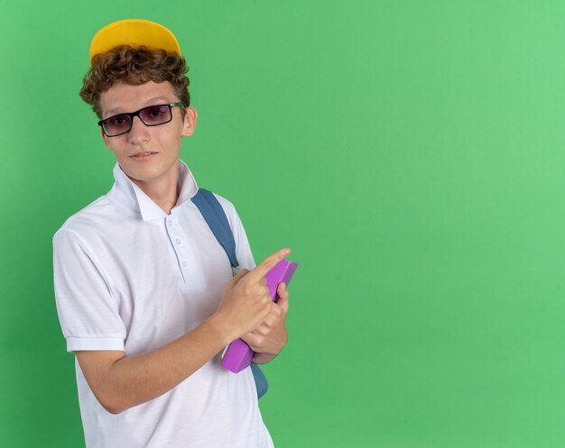Student guy in white shirt and yellow cap wearing glasses with backpack holding notebook looking at camera smiling confident pointing with index finger at copy space standing over green background