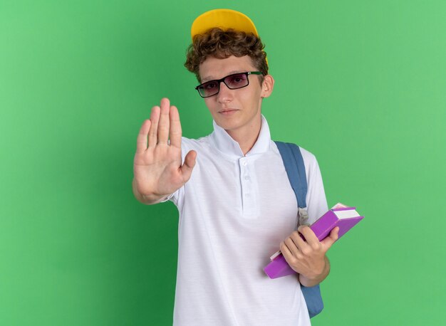 Student guy in white shirt and yellow cap wearing glasses with backpack holding notebooks looking at camera with serious face making stop gesture with hand