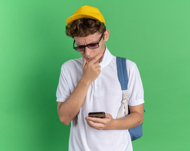 Student guy in white shirt and yellow cap wearing glasses with backpack holding smartphone looking at it puzzled standing over green background