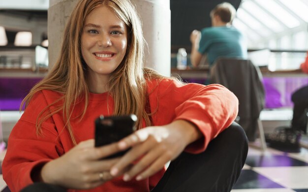 Студенческий образ жизни подростков и концепция городской жизни Улыбающаяся счастливая привлекательная рыжая девушка сидит на полу в кафе или коворкинге, отправляя текстовые сообщения друзьям с помощью мобильного телефона, играя на смартфоне