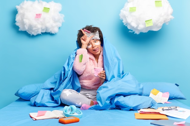 student makes loser gesture sticks out tongue wrapped in blanket studies at home poses on bed has coffee break