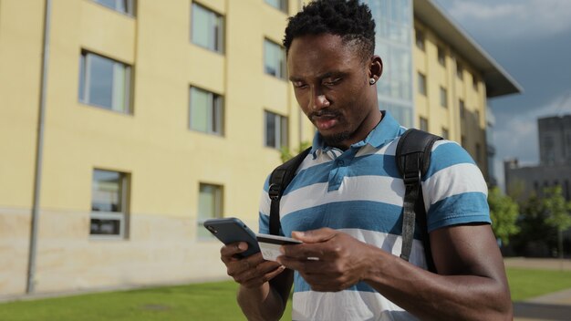 Студент-мужчина покупает и оплачивает в интернете на мобильном телефоне афро-американский мужчина, стоящий на улице и совершающий покупки в интернете с помощью кредитной карты на смартфоне