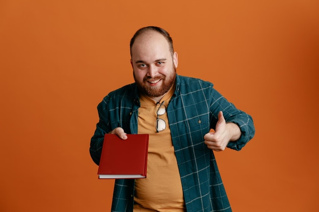 Student man in casual clothes holding notebook looking at camera happy and positive smiling showing thumb up standing over orange background