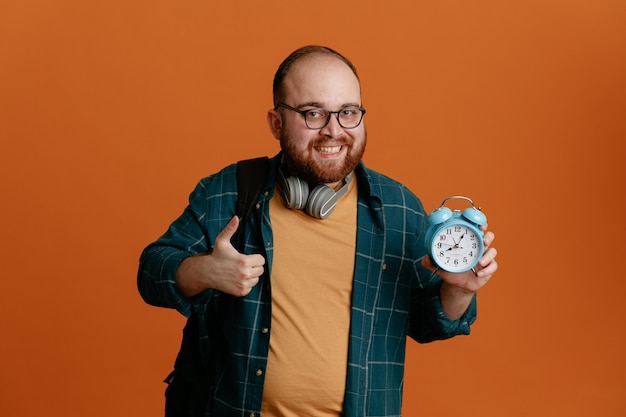 Student man in casual clothes wearing glasses with headphones holding alarm clock looking at camera happy and positive smiling cheerfully showing thumb up standing over orange background