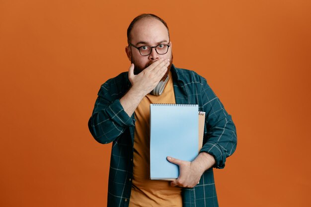 Student man in casual clothes wearing glasses with headphones holding notebooks looking at camera being amazed and shocked covering mouth with hand standing over orange background