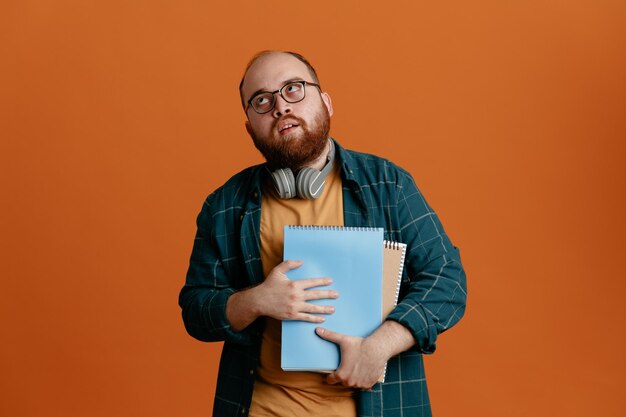 オレンジ色の背景の上に立って疲れて退屈して目を転がしているノートブックを保持しているヘッドフォンで眼鏡をかけているカジュアルな服を着た学生の男