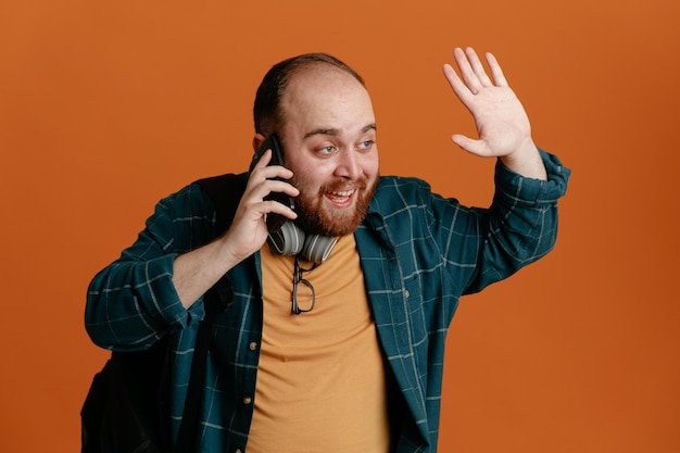 Student man in casual clothes with backpack with headphones talking on mobile phone happy and positive waving with hand standing over orange background