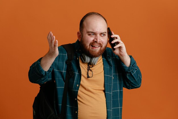 Student man in casual clothes with backpack with headphones talking on mobile phone looking annoyed and disappointed standing over orange background