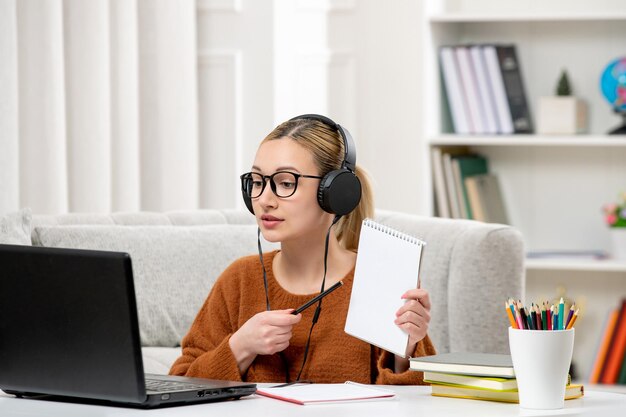 Student online cute girl in glasses and sweater studying on computer showing notes