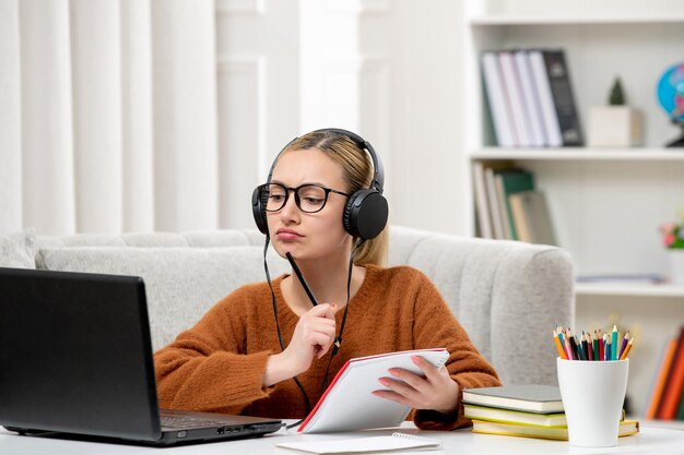 Студент онлайн милая девушка в очках и свитере учится на компьютере, думая ручкой и блокнотом