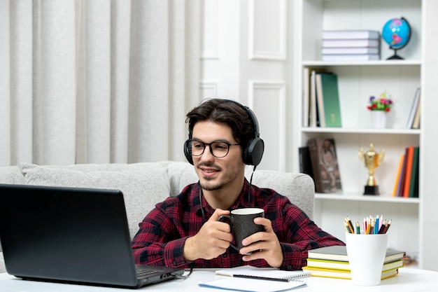 Студент онлайн симпатичный парень в клетчатой рубашке в очках учится на компьютере с чашкой кофе