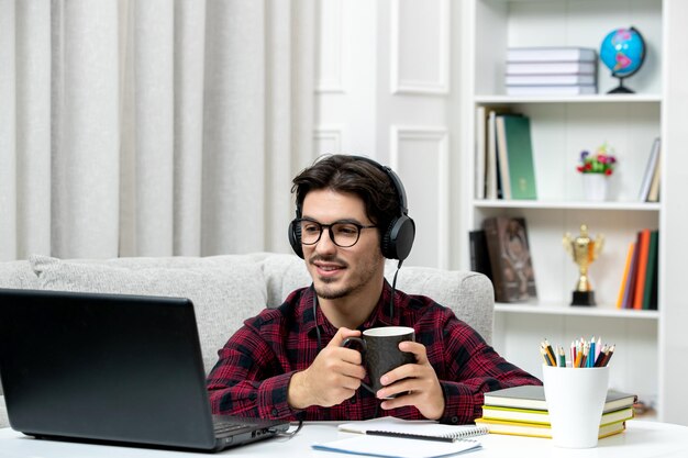 Student online cute guy in checked shirt with glasses studying on computer holding coffee cup