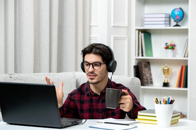 Student online cute guy in checked shirt with glasses studying on computer holding a cup