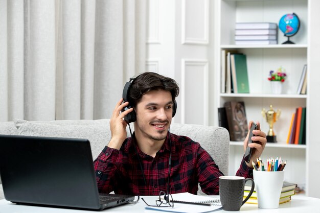 コンピューターの笑顔と電話で勉強している眼鏡をかけたチェックシャツの学生オンラインかわいい男