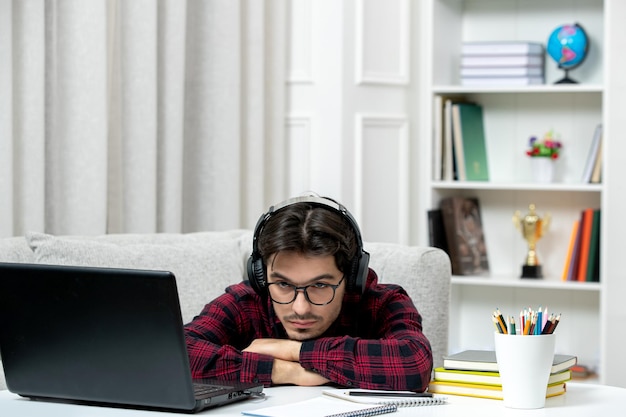 机の上に頭を置いてコンピューターで勉強している眼鏡をかけたチェックシャツの学生オンラインかわいい男