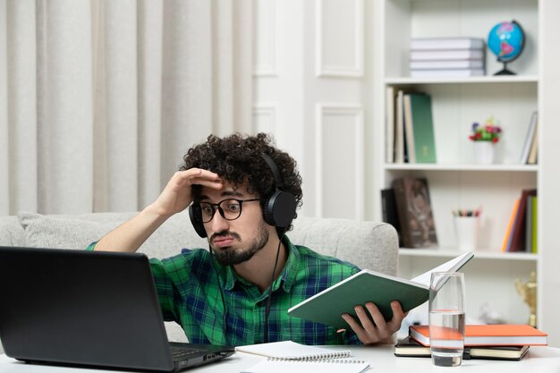 緑のシャツを着た眼鏡でコンピューターを勉強している学生のオンラインかわいい若い男は頭を抱えて混乱しました