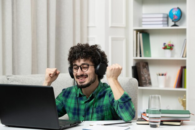 Student online cute young guy studying on computer in glasses in green shirt holding fists up