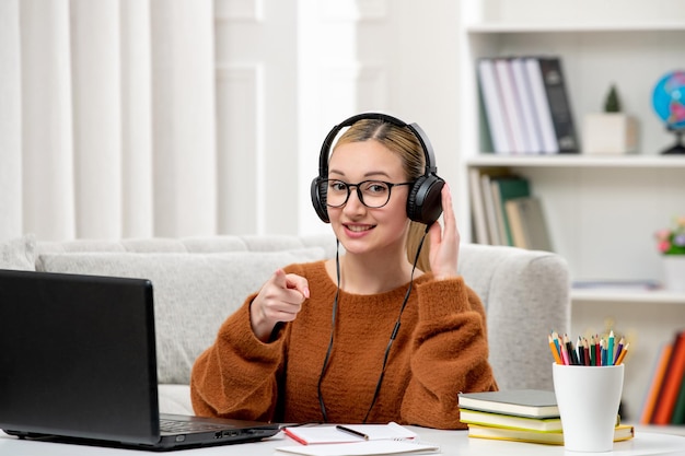 Studente online giovane ragazza carina con gli occhiali e maglione arancione che studia sul computer che punta in avanti