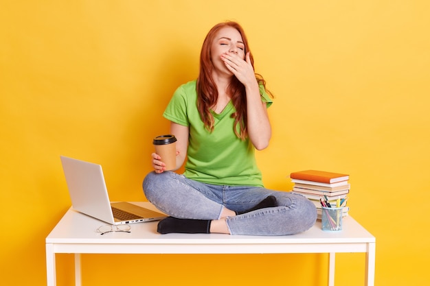 Student studying or preparing for exams, being tired and sleepy, sitting on table with crossed legs and yawning, wearing jeans and green t shirt isolated over yellow background.