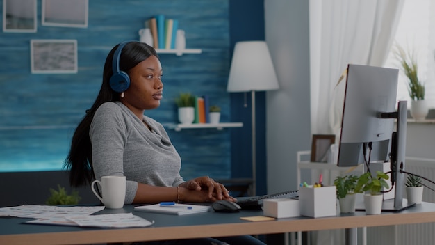 Student with black skin having headphone puts listening online univeristy course using elearning platform sitting at desk in living room