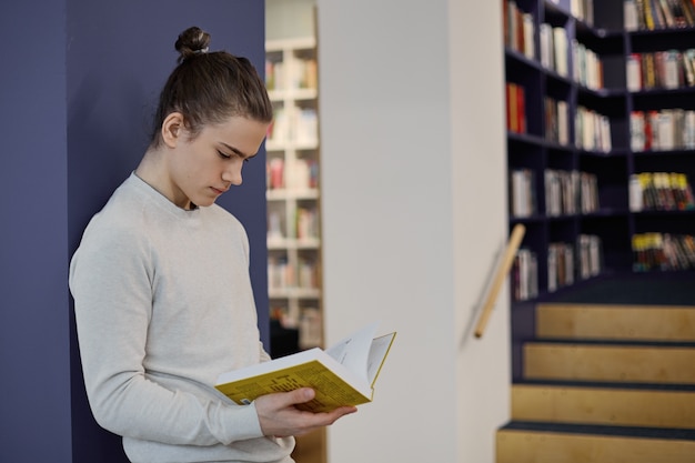 Student with hair knot standing in library, isolated reading information in open textbook on his hands while making research