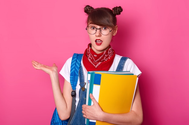 student with two bunches, wears t shirt, overalls, bandana, holds paper folder, takes palm aside, has astonished facial expression, poses with open mouth isolated over pink.