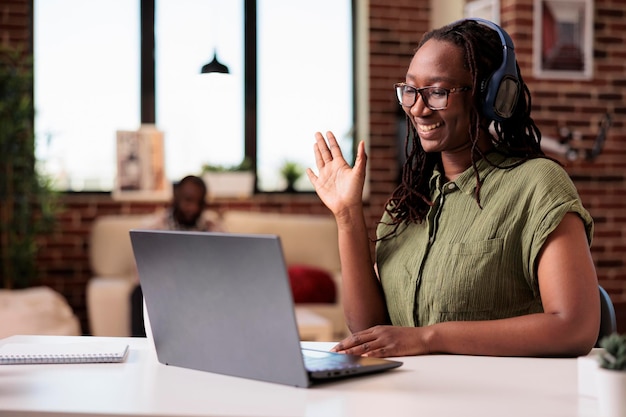 Free photo student with wireless headphones in video conference waving hello at colleagues while sitting at desk in living room. smiling freelancer greeting client in internet call while roommate is relaxing.