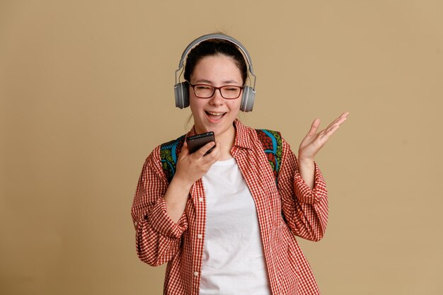 茶色の背景の上に幸せで肯定的な音声メッセージを録音する携帯電話を保持している彼女の頭の上のバックパックとヘッドフォンと眼鏡をかけているカジュアルな服装の学生の若い女性